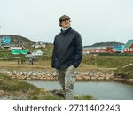 A man with a rain jacket and a flat cap, standing alone in Paamiut, a small town in Greenland. A few colorful houses behind him, and a small lake.