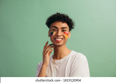 Man With Rainbow Face Paint Celebrates Gay Pride. Gay Man Standing On Grey Background Looking At Camera And Smiling.