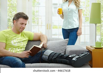 Man Rasting His Broken Leg In Cast On Sofa At Home, Reading Book.