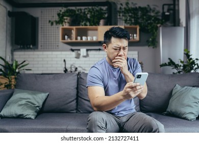 Man Reading Bad News From Phone, Asian Frustrated And Sad Looking At Phone Screen Sitting On Sofa At Home