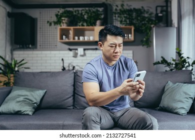 Man Reading Bad News From Phone, Asian Frustrated And Sad Looking At Phone Screen Sitting On Sofa At Home