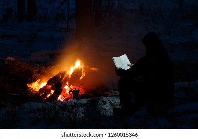 Man Reading A Book By The Fire. Book Is Highlighted By Headlamp Winter Night In The Forest
