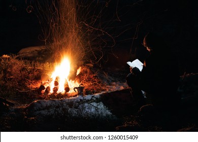 Man Reading A Book By The Fire. Book Is Highlighted By Headlamp Winter Night In The Forest