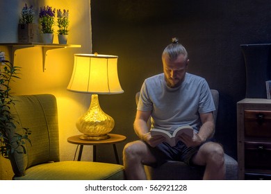 Man Reading A Book At Night. Young Man Sitting In The Living Room In The Twilight And Reading A Book. Horizontal Indoors Shot. 