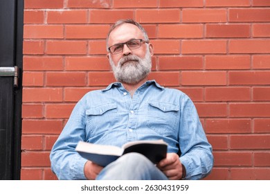 Man reading a book while sitting against a brick wall in a casual setting during a sunny afternoon in an urban area