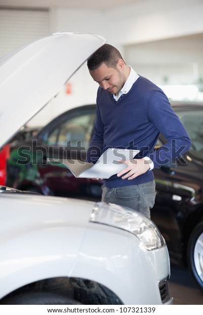 Man reading a file while looking at the engine in\
a garage