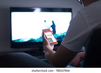 Man Reading Online News On Smartphone At Home. Mobile Phone News Website, Application Or Portal On Cellphone Screen. Holding Smart Device In Hand. Sitting In Living Room Late At Night.