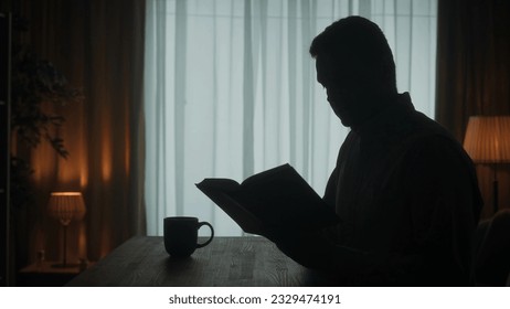 A man reads a book sitting at a table with a cup of coffee, tea in the room on the background of the window. Dark silhouette of a man with an open book in his hand close up. - Powered by Shutterstock