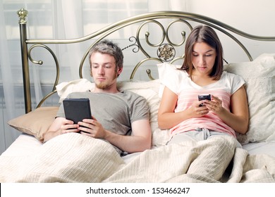 A man reads an e-book while his wife plays on a smartphone in their bedroom - Powered by Shutterstock