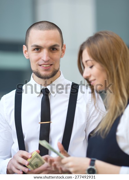 Man receiving money, US dollars. Business man\
holding a money hundred dollar banknotes. Businessman and\
businesswoman on blurring background office building work. They\
count the money and divide.