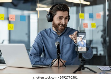 Man recording podcast in modern office setting with headphones, microphone, and smartphone. Professional setup for audio and video content creation. Engaging in remote communication, social media - Powered by Shutterstock