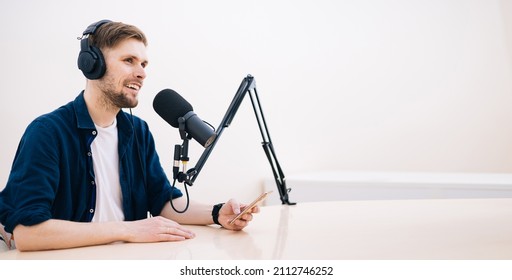 Man recording podcast in white studio. hand holding mobile phone in audio home studio. Male with headphones doing online meeting. Remote radio host broadcasting from home with professional microphone.