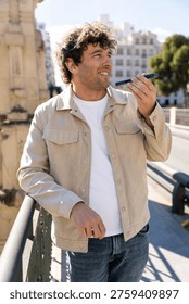 Man recording a voice message using a smartphone, smiling happily in an urban setting, enjoying modern communication