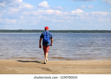 A Man In A Red Baseball Cap And A Blue Plaid Shirt Walks Along The Shore Of The Reservoir.