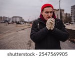 Man in red beanie and gloves standing outdoors in cold weather near urban area.