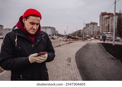 Man in a red beanie stands on a sidewalk looking at his smartphone in an urban setting during overcast weather. - Powered by Shutterstock