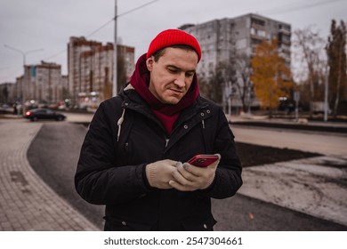 Man in a red beanie stands on a sidewalk looking at his smartphone in an urban setting during overcast weather. - Powered by Shutterstock