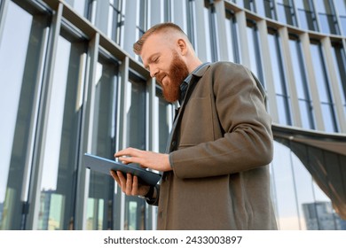 A man with a red beard dressed in a shirt and jacket checks his email on a tablet. - Powered by Shutterstock