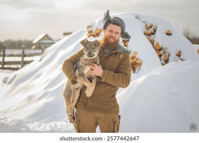 A man with a red beard in a hat with earflaps in winter holds a dog in his arms against the backdrop of snow and the village.
