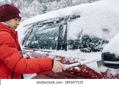 The Man In Red Coat Cleans The Car With Brush During Snowfall. Bad Snow Weather Concept. Winter Inclement Weather.