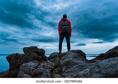 A man in a red jacket with a backpack on his back stands on the sea rocks and looks at the sea against the backdrop of storm clouds