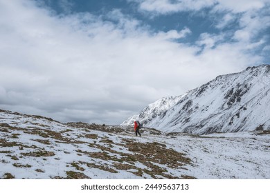Man in red jacket with big backpack walks through grassy rocky pass covered snow. Backpacker goes on freshly fallen snow in high mountains in changeable weather. Snow covered stony hill with grass.