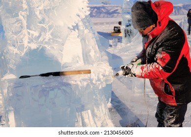 A Man In A Red Jacket Cuts A Sculpture Out Of A Piece Of Transparent Ice With A Special Tool - An Electric Jigsaw. Sunny Day In Winter. Ice Sculpture Festival