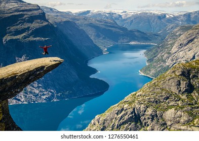 A man in a red jacket jumping on the Trolltunga rock with a blue lake 700 meters lower and interesting sky with clouds