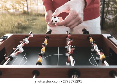 A man in a red jacket playing table foosball in a garden, holding a small soccer ball above a table with red and yellow players arranged on a miniature field, capturing a casual outdoor moment that shows play is ageless and brings joy in any season