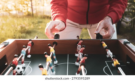 A man in a red jacket plays table foosball in a garden. The red and yellow teams stand on a green field, capturing a casual game that echoes the excitement and anticipation of major world football events, including upcoming global soccer championship