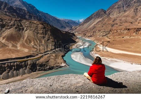 Man in red jacket sit on cliff and look at Sangam or confluence of Indus and Zanskar Rivers at Leh, Ladakh, India.