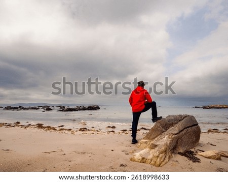A man in a red jacket is standing by a rock on a sandy beach. The sky is cloudy and the beach is empty. Cloudy sky. West coast of Ireland. Travel and tourism. Outdoor activity
