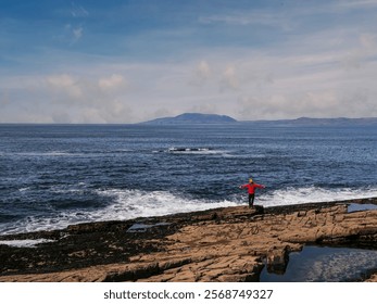 A man in a red jacket stands on a rocky beach looking out at the ocean. The sky is clear and the water is calm. Mullaghmore peninsula Ireland. Travel and tourism. Trip to Irish nature - Powered by Shutterstock