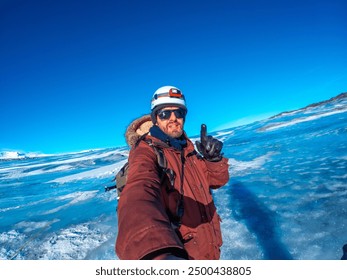 A man in a red jacket is taking a selfie in front of a blue body of water. He is wearing a helmet and gloves, and he is smiling - Powered by Shutterstock