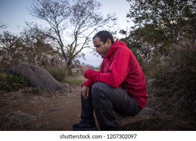 Man in red jacket using phone in left hand beside dirt trail in nature