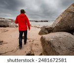 A man in a red jacket walks on a beach near a large rock. The sky is cloudy and the beach is empty. Cloudy sky. West coast of Ireland. Travel and tourism. Outdoor activity