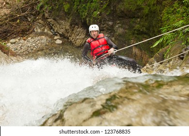 A man in a red life jacket and white helmet rappels down a powerful waterfall gripping a rope in a rocky forested canyon in Banos, Ecuador, splashing white water and mossy banks framing the adventure.