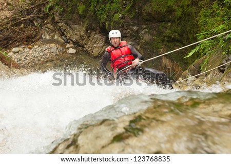 A man in a red life jacket and white helmet rappels down a powerful waterfall gripping a rope in a rocky forested canyon in Banos, Ecuador, splashing white water and mossy banks framing the adventure.