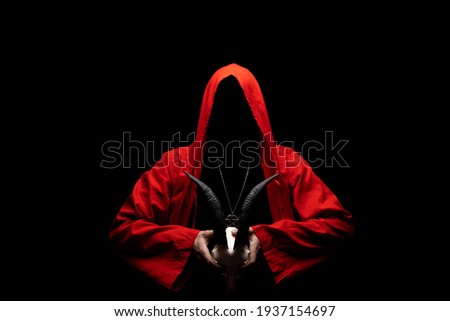 Man in red ritual hooded cloak holds a skull with horns in hands. Religious sects, satanism concept. No face. 