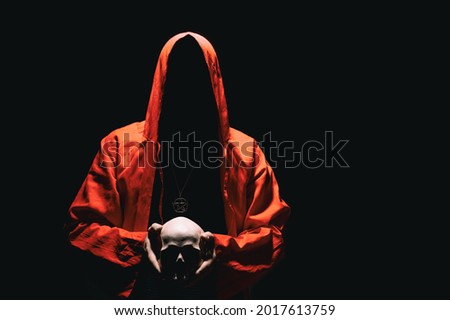 Man in red ritual hooded cloak holds a human skull in hands. Religious sects, satanism, occult, esoteric, concept. Copy space.