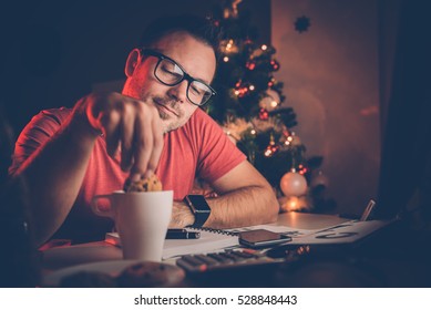 Man in red shirt dipping a cookie in coffee at home office with christmas tree behind him