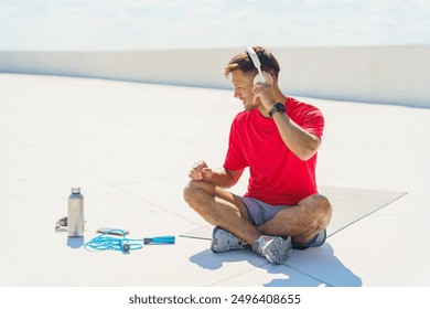 Man in a red shirt sits on a mat outdoors, adjusting his headphones and preparing for a workout.

 - Powered by Shutterstock