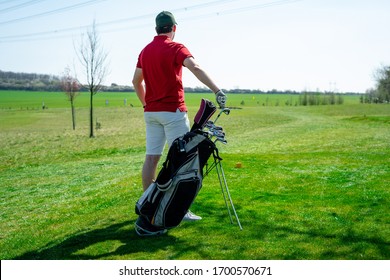 Man in red shirt, white shorts and baseball cap preparing to hit golf ball with club. He has golf clubs next to him. Sports that people around the world play during the quarantine for health.