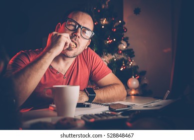 Man in red shirt working late and eating cookie with christmas tree behind him