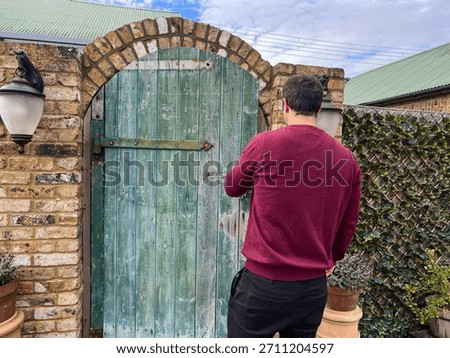 Man in red sweater closing rustic green wooden gate in brick courtyard garden