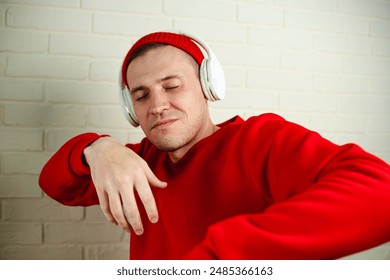 A man in a red sweatshirt dances while listening to music through headphones. He is standing in front of a white brick wall, and his eyes are closed as he enjoys the music