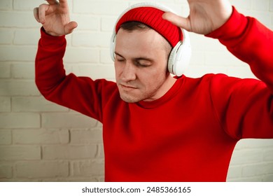 A man in a red sweatshirt dances while listening to music through headphones. He is standing in front of a white brick wall, and his eyes are closed as he enjoys the music
