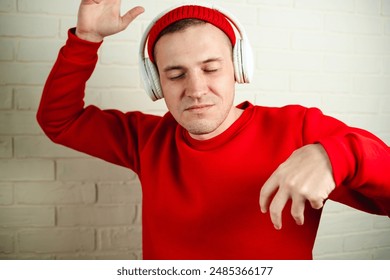 A man in a red sweatshirt dances while listening to music through headphones. He is standing in front of a white brick wall, and his eyes are closed as he enjoys the music