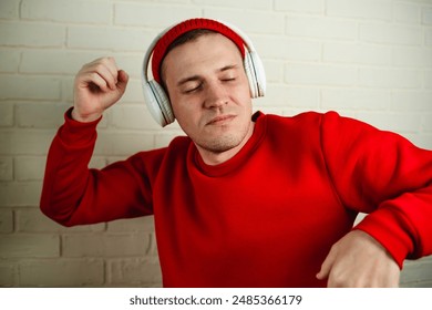 A man in a red sweatshirt dances while listening to music through headphones. He is standing in front of a white brick wall, and his eyes are closed as he enjoys the music