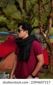 A man in red wearing sunglasses and a black scarf is looking outside against a blurry background of nature and a tent.  Handsome Asian male model. - Shutterstock ID 2394607117